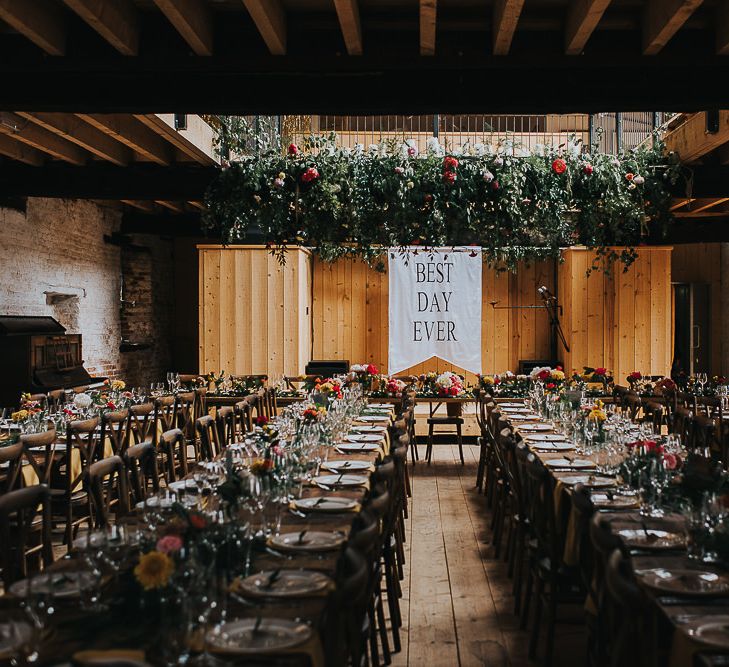 Best Day Ever Flag | Hanging Floral Installation | Long Wooden Banquet Tables | Yellow Napkins | Pink, Orange and Yellow Table Flowers with Foliage | Wedding Reception at Abbey Hall Creative Space | 'Frida Kahlo' Flower Crown, Halfpenny London Bridal Separates and Colourful Bouquets with Peonies for Suffolk Wedding | From The Smiths Photography