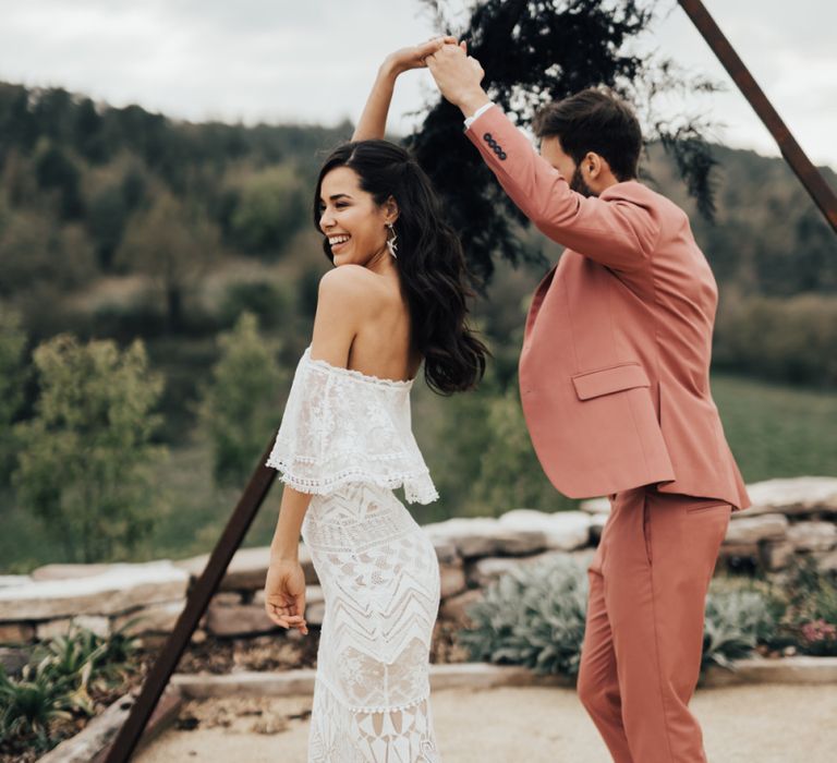 Groom in Pink Suit Twirling  His Bride in Grace Loves Lace Wedding Dress in Front of a Wooden Triangle Structure