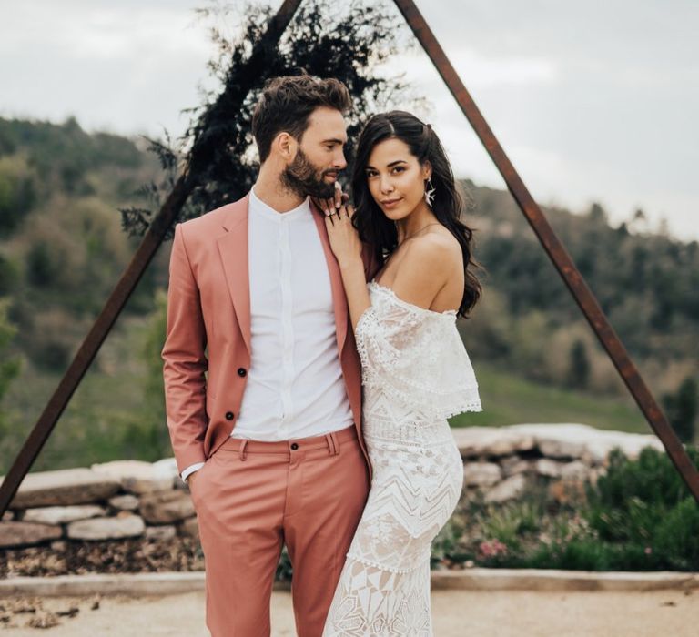 Boho Bride in Bardot Grace Loves Lace Wedding Dress and Groom in Coral Suit Standing in Front of a Wooden Triangle Altar