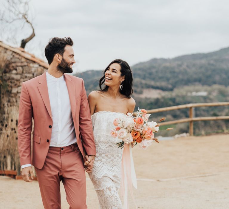 Boho Bride in Bardot Grace Loves Lace Wedding Dress and Groom in Coral Suit  Arm in Arm