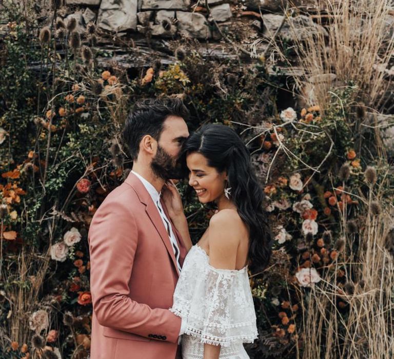 Bride in Off The Shoulder Lace Wedding Dress and Groom in Dusky Pink Suit Laughing by a Dried Flower Arrangement