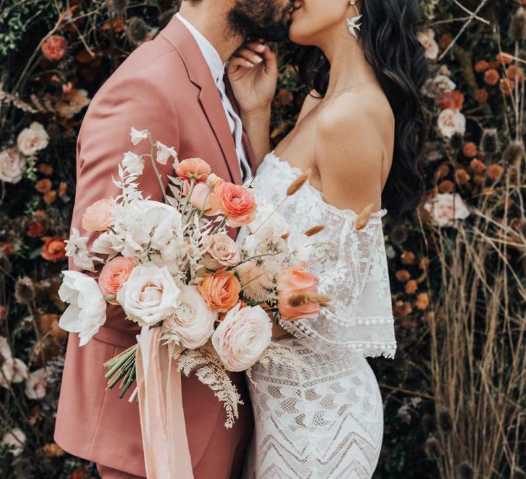 Bride in Off The Shoulder Lace Wedding Dress and Groom in Dusky Pink Suit  Kissing, Holding a White and Peach Wedding Bouquet