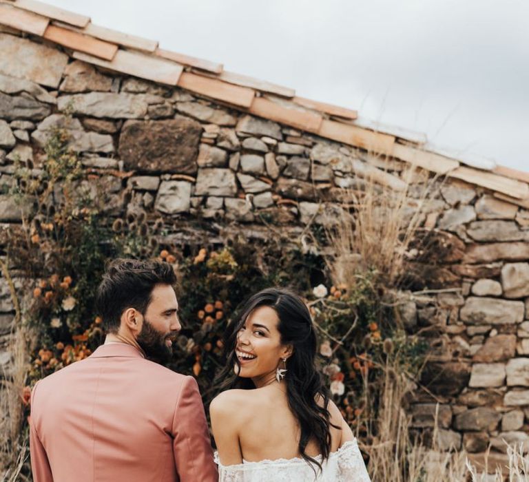 Stylish Bride in Grace Loves Lace Wedding Dress and Groom in Dusky Pink Suit Holding Hands Laughing