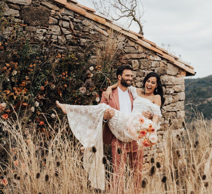 Groom in Coral Wedding Suit Picking Up His Bride in a Grace Loves Lace Wedding Dress