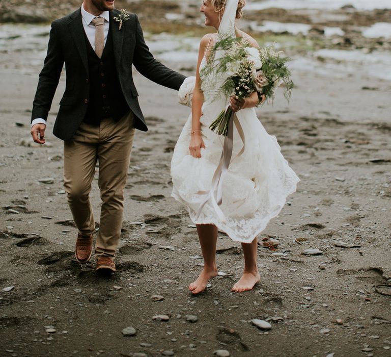 Bride in lace Willowby by Watters wedding dress and groom in chino's and blazer walking on the beach