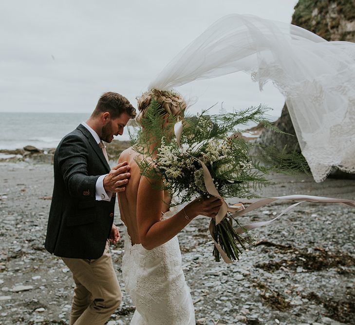 Bride in lace Willowby by Watters wedding dress and fly away veil and groom in chino's and blazer walking on the beach