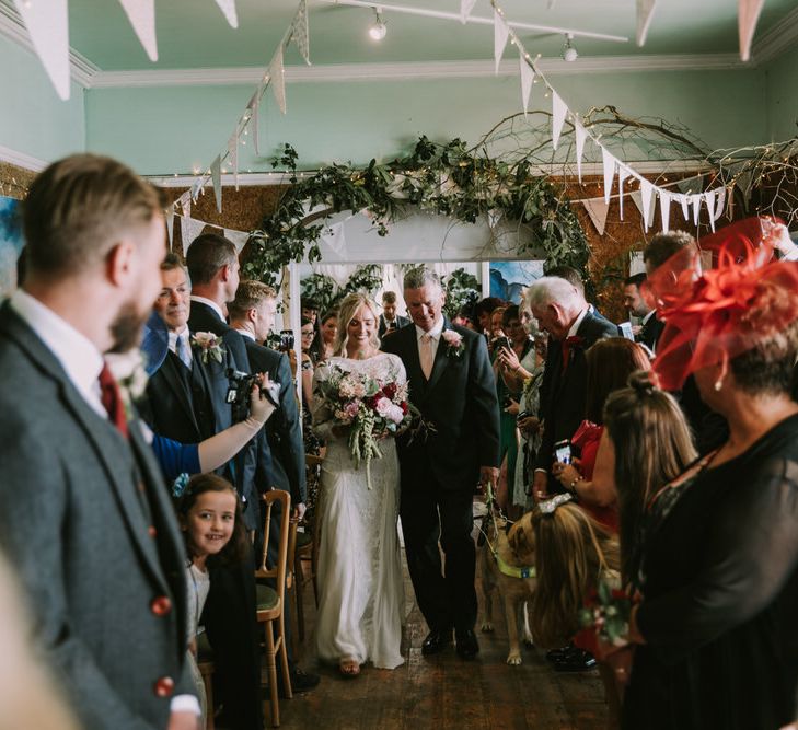 Bride in Grace Loves Lace Inca Gown with Boat Neck, Lace Sleeves and Low Back | Bridal Bouquet of Burgundy and Blush Flowers with Foliage | Groom in Three-Piece Grey Herringbone Suit with Burgundy Tie and Buttons | Father of the Bride in Grey Three-Piece Suit with Blush Tie | Foliage Arch | Pastel Bunting | Fairy Lights | Jackson the Guide Dog | Wedding Ceremony at The Druidstone | Mykonos Rewritten Bridesmaid Dresses for an Epic Clifftop Coastal Wedding | Nic Ford Photography