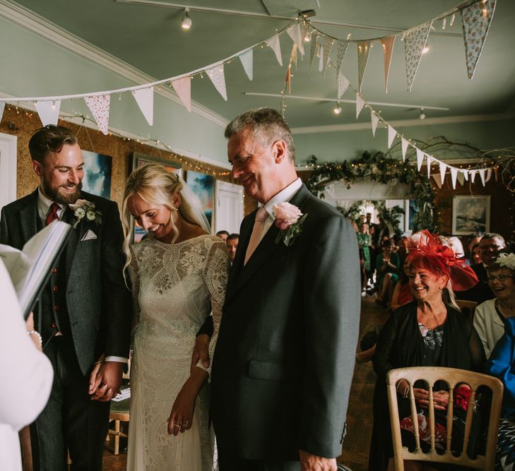 Bride in Grace Loves Lace Inca Gown with Boat Neck, Lace Sleeves and Low Back | Groom in Three-Piece Grey Herringbone Suit with Burgundy Tie and Buttons | Blush Buttonhole | Father of the Bride in Grey Three-Piece Suit with Blush Tie | Foliage Arch | Pastel Bunting | Fairy Lights | Wedding Ceremony at The Druidstone | Mykonos Rewritten Bridesmaid Dresses for an Epic Clifftop Coastal Wedding | Nic Ford Photography