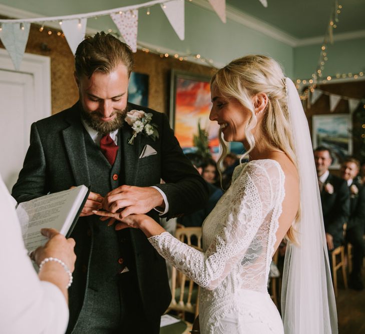 Bride in Grace Loves Lace Inca Gown with Boat Neck, Lace Sleeves and Low Back | Personalised Embroidered Veil by Daisy Sheldon | Aurora Hair Comb by Tilly Thomas Lux  | Groom in Three-Piece Grey Herringbone Suit with Burgundy Tie and Buttons | Blush Buttonhole | Grey Pocket Square | Pastel Bunting | Fairy Lights | Wedding Ceremony at The Druidstone | Mykonos Rewritten Bridesmaid Dresses for an Epic Clifftop Coastal Wedding | Nic Ford Photography