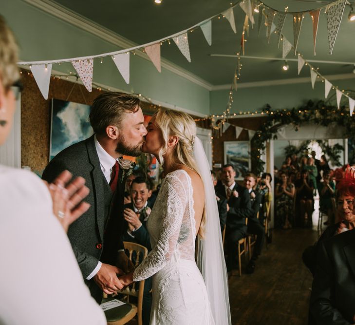 Bride in Grace Loves Lace Inca Gown with Boat Neck, Lace Sleeves and Low Back | Personalised Embroidered Veil by Daisy Sheldon | Aurora Hair Comb by Tilly Thomas Lux  | Groom in Three-Piece Grey Herringbone Suit with Burgundy Tie and Buttons | Blush Buttonhole | Foliage Arch | Pastel Bunting | Fairy Lights | Wedding Ceremony at The Druidstone | Mykonos Rewritten Bridesmaid Dresses for an Epic Clifftop Coastal Wedding | Nic Ford Photography