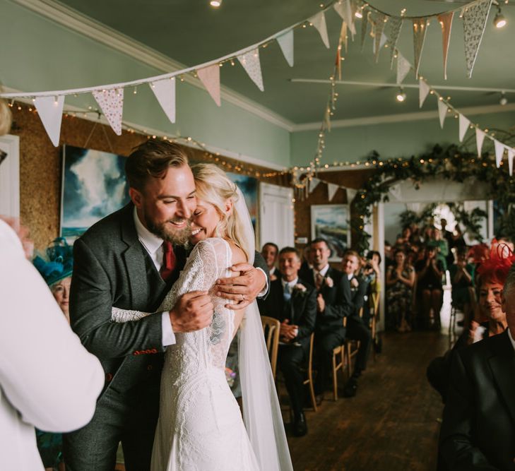 Bride in Grace Loves Lace Inca Gown with Boat Neck, Lace Sleeves and Low Back | Personalised Embroidered Veil by Daisy Sheldon | Groom in Three-Piece Grey Herringbone Suit with Burgundy Tie and Buttons | Foliage Arch | Pastel Bunting | Fairy Lights | Wedding Ceremony at The Druidstone | Mykonos Rewritten Bridesmaid Dresses for an Epic Clifftop Coastal Wedding | Nic Ford Photography