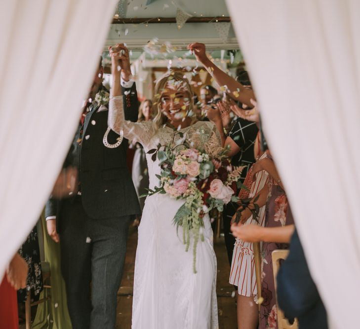 Bride in Grace Loves Lace Inca Gown with Boat Neck, Lace Sleeves and Low Back | Bridal Bouquet of Burgundy and Blush Flowers with Foliage | Groom in Three-Piece Grey Herringbone Suit with Burgundy Tie and Buttons | Pastel Confetti | Wedding Ceremony at The Druidstone | Mykonos Rewritten Bridesmaid Dresses for an Epic Clifftop Coastal Wedding | Nic Ford Photography