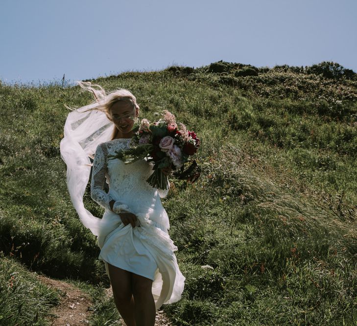 Bride in Grace Loves Lace Inca Gown with Boat Neck, Lace Sleeves and Low Back | Bridal Bouquet of Burgundy and Blush Flowers with Foliage | Mykonos Rewritten Bridesmaid Dresses for an Epic Clifftop Coastal Wedding | Nic Ford Photography