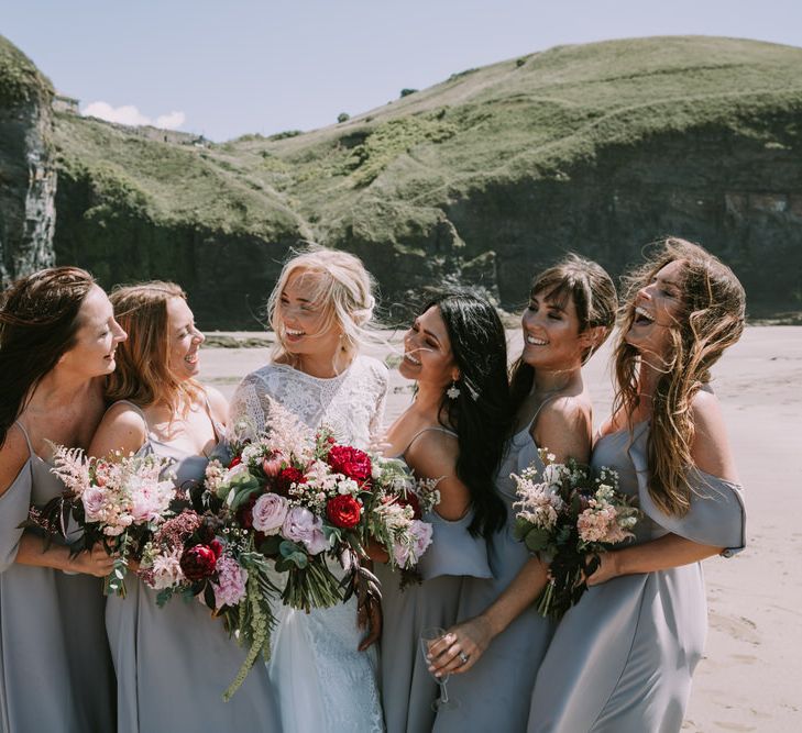 Bride in Grace Loves Lace Inca Gown with Boat Neck, Lace Sleeves and Low Back | Bridesmaids in Grey Cold Shoulder Dresses by Rewritten | Bridal Party Bouquets of Burgundy and Blush Flowers with Foliage | Mykonos Rewritten Bridesmaid Dresses for an Epic Clifftop Coastal Wedding | Nic Ford Photography