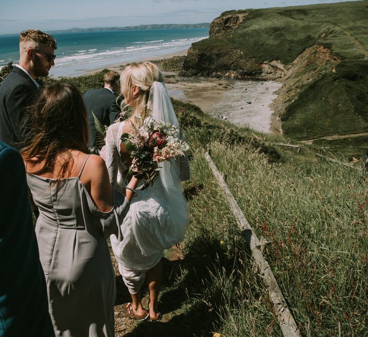 Bride in Grace Loves Lace Inca Gown with Boat Neck, Lace Sleeves and Low Back | Personalised Embroidered Veil by Daisy Sheldon | Aurora Hair Comb by Tilly Thomas Lux  | Bridesmaid in Grey Cold Shoulder Dress by Rewritten | Bouquet of Blush and Pink Flowers with Foliage | Mykonos Rewritten Bridesmaid Dresses for an Epic Clifftop Coastal Wedding | Nic Ford Photography