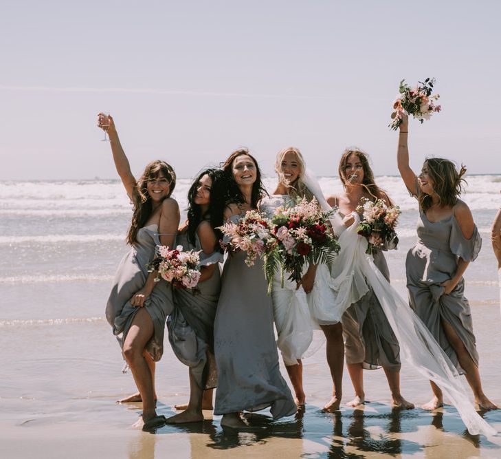 Bride in Grace Loves Lace Inca Gown with Boat Neck, Lace Sleeves and Low Back | Personalised Embroidered Veil by Daisy Sheldon | Bridesmaids in Grey Cold Shoulder Dresses by Rewritten | Bridal Party Bouquets of Burgundy and Blush Flowers with Foliage | Paddling in the Sea at Druidston Beach | Mykonos Rewritten Bridesmaid Dresses for an Epic Clifftop Coastal Wedding | Nic Ford Photography