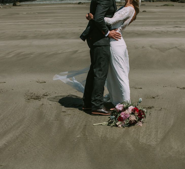 Bridal Bouquet of Burgundy and Blush Flowers with Foliage | Bride in Grace Loves Lace Inca Gown with Boat Neck, Lace Sleeves and Low Back | Groom in Three-Piece Grey Herringbone Suit with Burgundy Tie and Buttons | Druidston Beach  | Mykonos Rewritten Bridesmaid Dresses for an Epic Clifftop Coastal Wedding | Nic Ford Photography