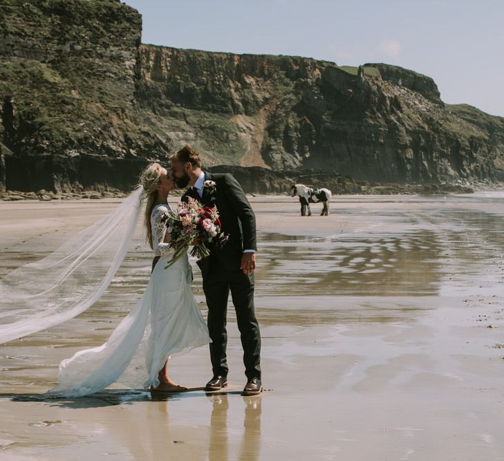 Bride in Grace Loves Lace Inca Gown with Boat Neck, Lace Sleeves and Low Back | Personalised Embroidered Veil by Daisy Sheldon | Bridal Bouquet of Burgundy and Blush Flowers with Foliage | Groom in Three-Piece Grey Herringbone Suit with Burgundy Tie and Buttons | Blush Buttonhole | Druidston Beach | Mykonos Rewritten Bridesmaid Dresses for an Epic Clifftop Coastal Wedding | Nic Ford Photography