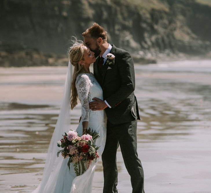 Bride in Grace Loves Lace Inca Gown with Boat Neck, Lace Sleeves and Low Back | Personalised Embroidered Veil by Daisy Sheldon | Bridal Bouquet of Burgundy and Blush Flowers with Foliage | Groom in Three-Piece Grey Herringbone Suit with Burgundy Tie and Buttons | Blush Buttonhole | Druidston Beach | Mykonos Rewritten Bridesmaid Dresses for an Epic Clifftop Coastal Wedding | Nic Ford Photography