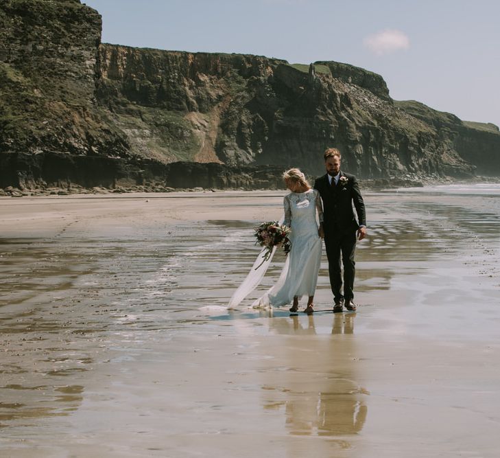 Bride in Grace Loves Lace Inca Gown with Boat Neck, Lace Sleeves and Low Back | Personalised Embroidered Veil by Daisy Sheldon | Bridal Bouquet of Burgundy and Blush Flowers with Foliage | Groom in Three-Piece Grey Herringbone Suit with Burgundy Tie and Buttons | Blush Buttonhole | Druidston Beach | Mykonos Rewritten Bridesmaid Dresses for an Epic Clifftop Coastal Wedding | Nic Ford Photography