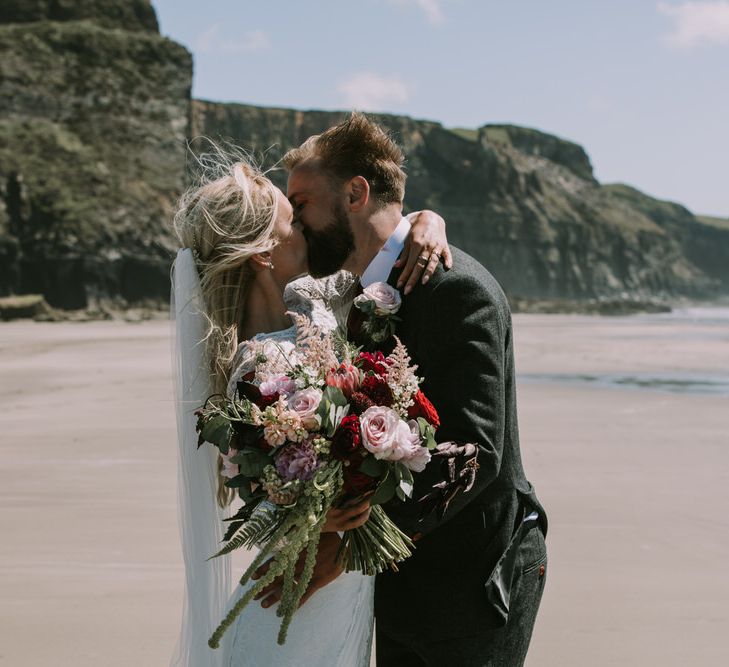 Bride in Grace Loves Lace Inca Gown with Boat Neck, Lace Sleeves and Low Back | Personalised Embroidered Veil by Daisy Sheldon | Aurora Hair Comb by Tilly Thomas Lux | Bridal Bouquet of Burgundy and Blush Flowers with Foliage | Groom in Three-Piece Grey Herringbone Suit with Burgundy Tie and Buttons | Blush Buttonhole | Druidston Beach | Mykonos Rewritten Bridesmaid Dresses for an Epic Clifftop Coastal Wedding | Nic Ford Photography