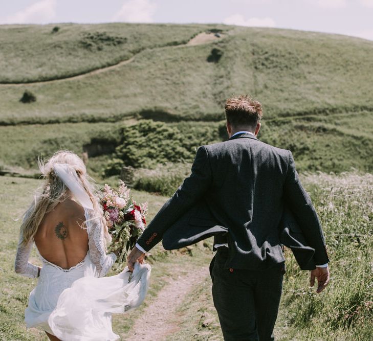 Bride in Grace Loves Lace Inca Gown with Boat Neck, Lace Sleeves and Low Back | Personalised Embroidered Veil by Daisy Sheldon | Aurora Hair Comb by Tilly Thomas Lux | Bridal Bouquet of Burgundy and Blush Flowers with Foliage | Groom in Three-Piece Grey Herringbone Suit with Burgundy Tie and Buttons | Druidston Beach | Mykonos Rewritten Bridesmaid Dresses for an Epic Clifftop Coastal Wedding | Nic Ford Photography