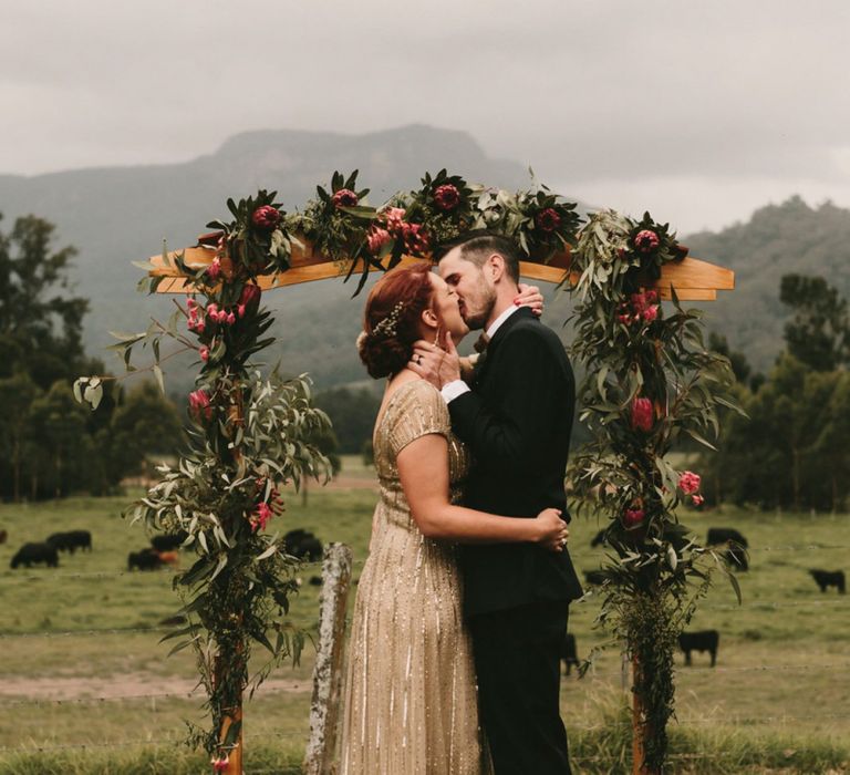 King Protea Floral Altar with Bride in Sequin Gold Wedding Dress and Groom  in Dark Suit Kissing