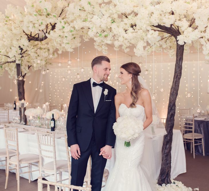 Bride in Beaded Fishtail Wedding Dress and Groom in Tuxedo Standing in Front of Blossom Tress