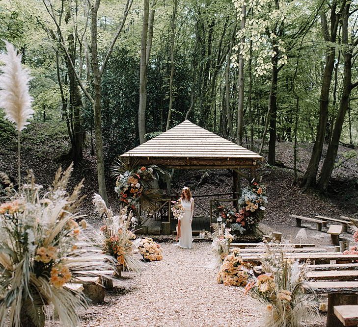 Outdoor Woodland Ceremony with Wooden Benches, and Hut Decorated in Floral Arrangements