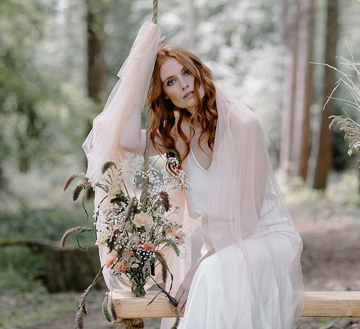 Beautiful Bride in a Fitted Wedding Dress and Blush Tulle Stole Sitting on a Swing in the Woodland