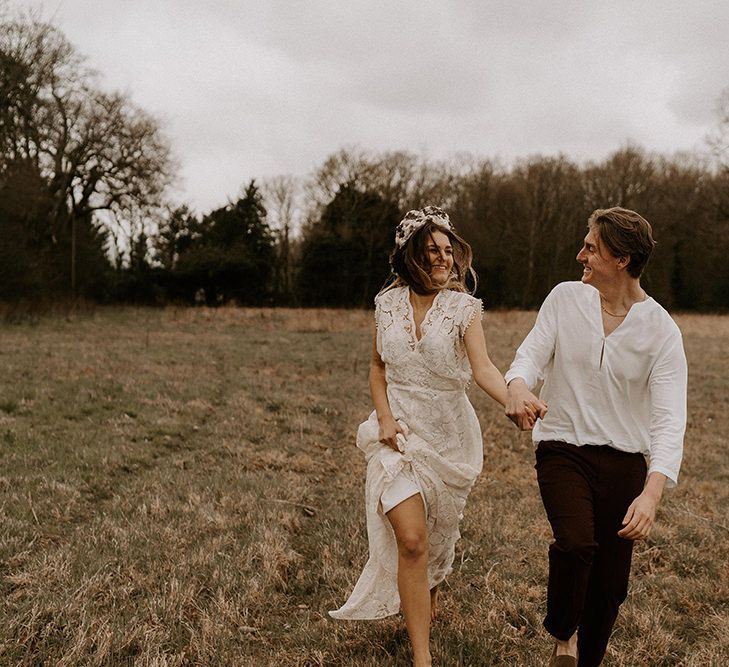 Boho Bride in Lace Wedding Dress and Groom in Casual Trousers and Shirt Running Through The Fields