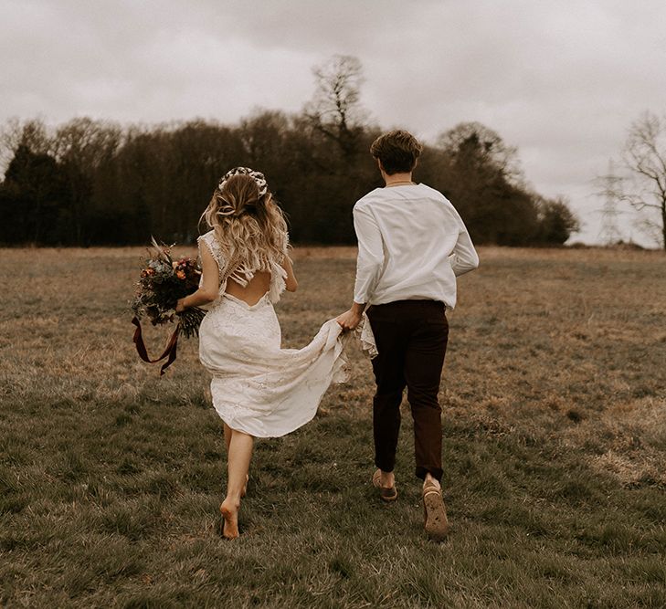 Boho Bride in Lace Wedding Dress and Groom in Casual Shirt and Trousers Running Through the Field