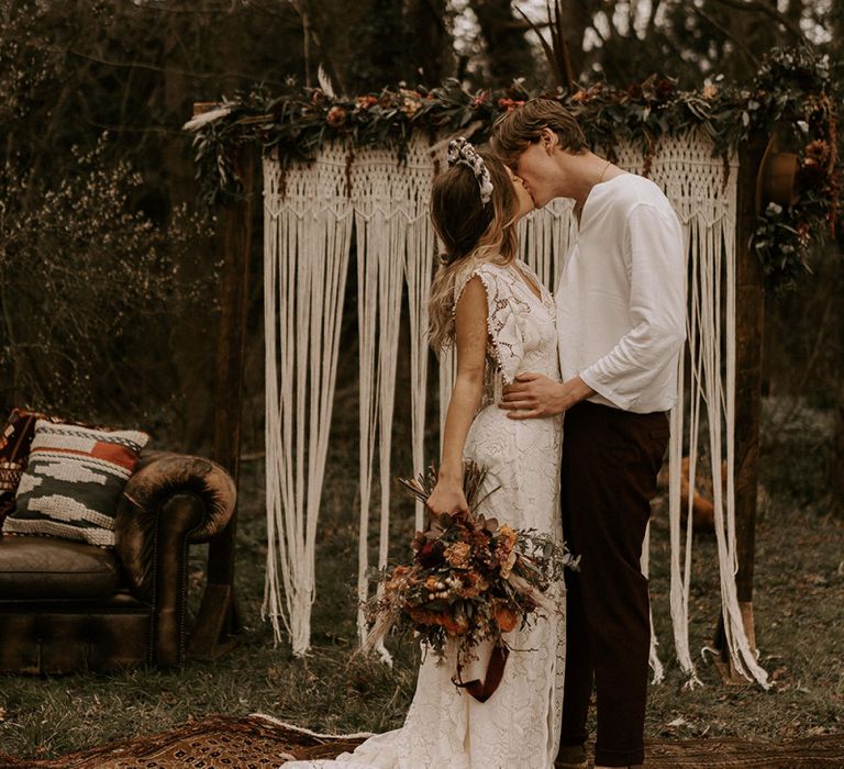 Boho Bride in Lace Wedding Dress Kissing Her Groom in Casual Shirt and Trousers at the Altar of Macrame Backdrop and Persian Rug Floor