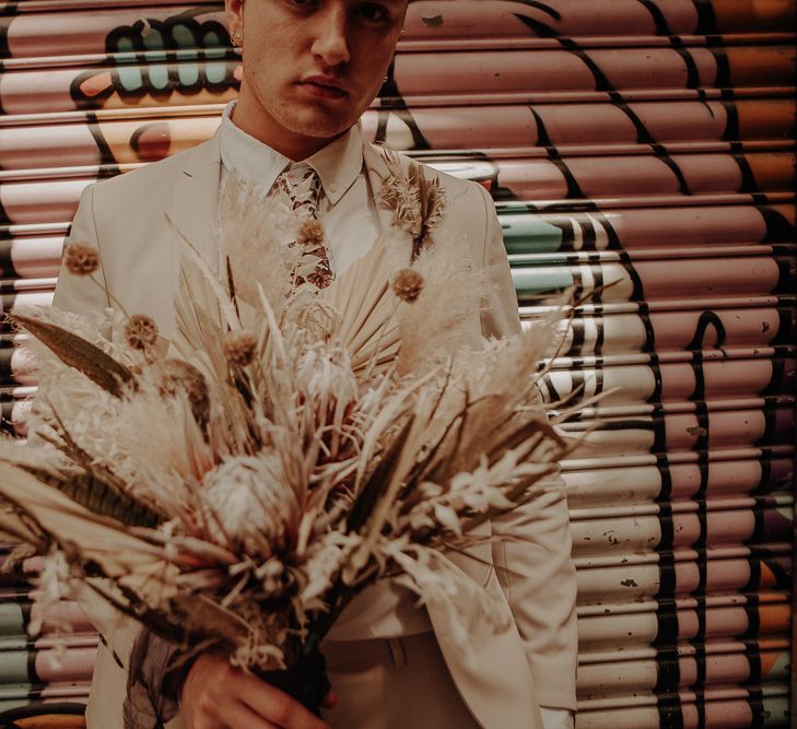 Groom in beige suit with floral tie holding a dried flower bouquet