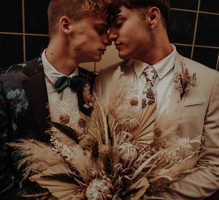 Grooms embracing in patterned and beige suits holding a bouquet with dried grasses and flowers