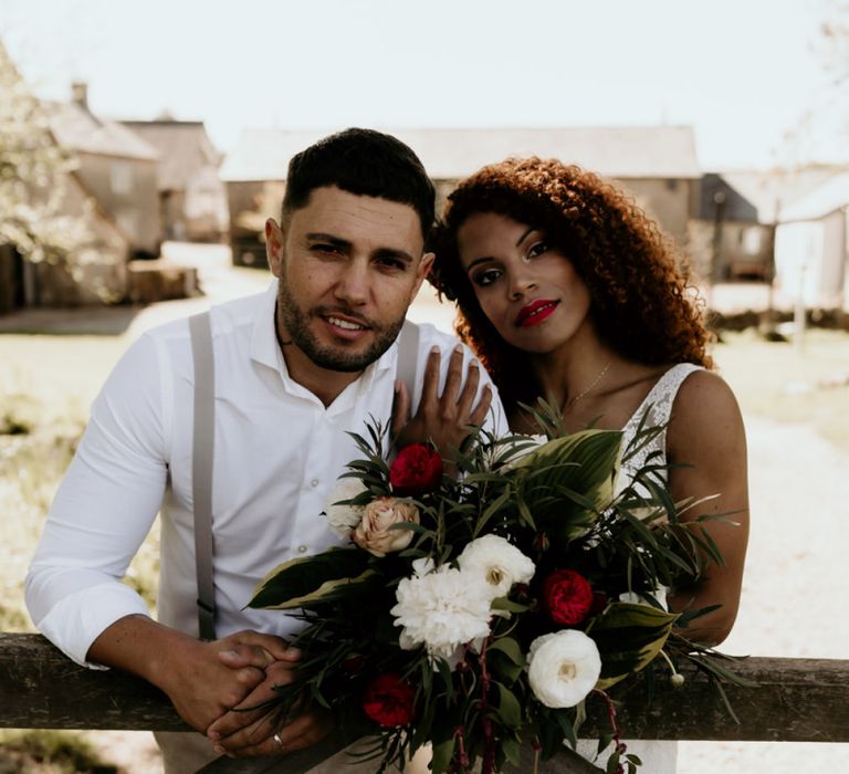 Black Bride in Lace Wedding Dress and Groom in White Shirt and Braces Embracing Next to Country Fence