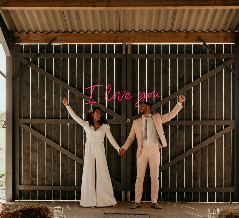 Black Bride with Afro Hair  in White Jumpsuit with Groom in Pink Suit Standing Next to Neon I Love You Sign