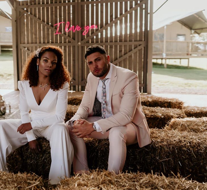 Black Bride with Afro Hair in Jumpsuit and Groom in Pink Suit Sitting On Hay Bales in a Barn