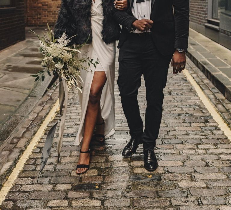 Stylish black bride in satin wedding dress and faux fur cover up and groom in bow tie and hat