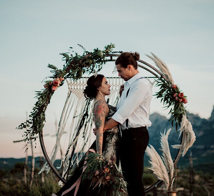 Alternative Bride and Groom Standing in Front of Steel Moon Gate Decorated with Macrame, Red Flowers, Foliage and Pampas Grass