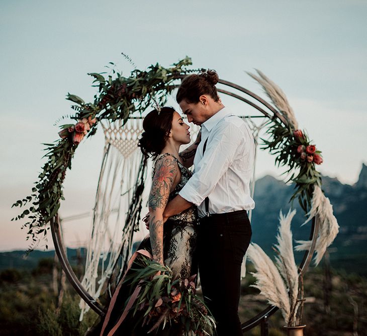 Bride and Groom Standing in Front of Steel Moon Gate Decorated with Macrame, Red Flowers, Foliage and Pampas Grass