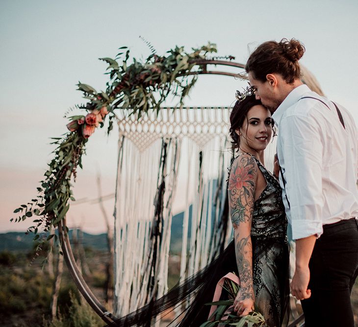 Bride in Antonia Serena Atelier Wedding Dress with Groom Standing in Front of Steel Moon Gate Decorated with Macrame, Red Flowers, Foliage and Pampas Grass