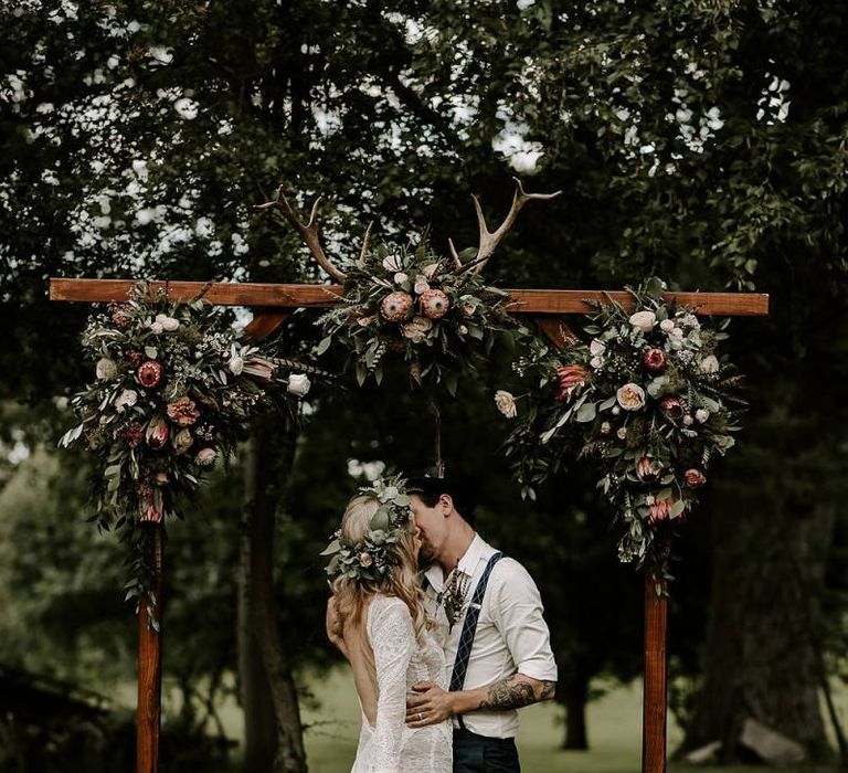 Bride clutches Protea bouquet as she kisses groom under wooden arch decorated in flowers