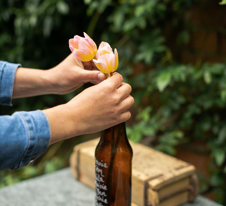 Person putting two pink and yellow tulips inside a brown beer bottle to create a wedding seating plan