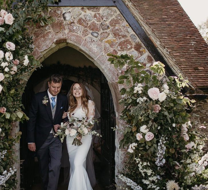 Bride in Lace St Partrick Wedding Dress and Groom in Morning Suit Exiting the Church Wedding Ceremony with Doorway Floral Arrangements