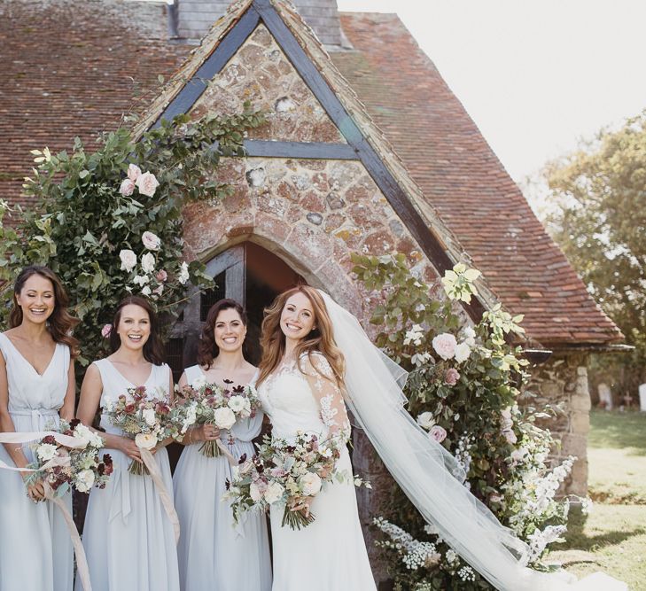 Bridal Party Standing in Front of the Church Entrance with Bridesmaids in Grey Dresses and Bride in Lace Wedding Dress