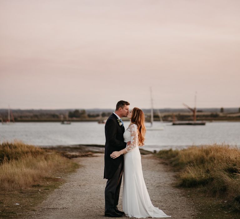 Bride in St Patrick Wedding Dress and Groom in Traditional Morning Suit Embracing by the Swale estuary