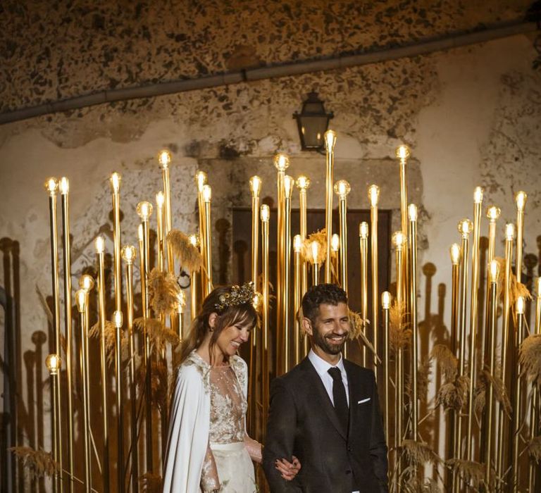 bride walking with groom wearing a bridal cape and gold bridal crown