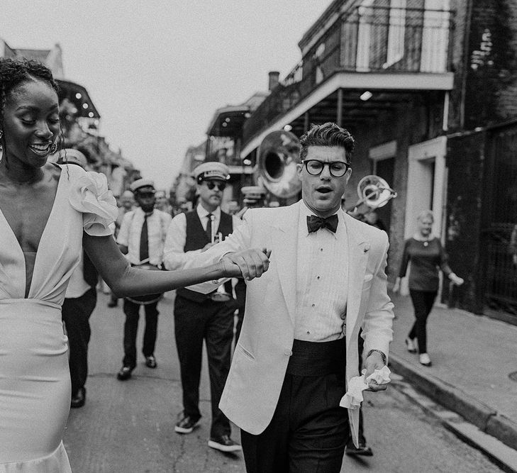 Stylish Bride and Groom in Johanna Ortiz  Wedding Dress and White Tuxedo Jacket Walking Down the Street with a Brass Band