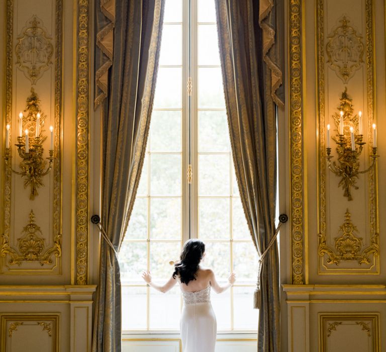 Bride Standing at a Grand Window Showing off the Back of Her Tara Keely Wedding Dress